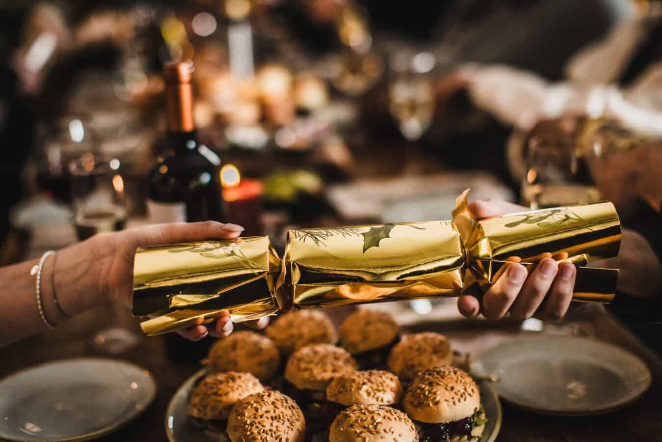 Hands holding a gold-wrapped Christmas cracker at a festive table.