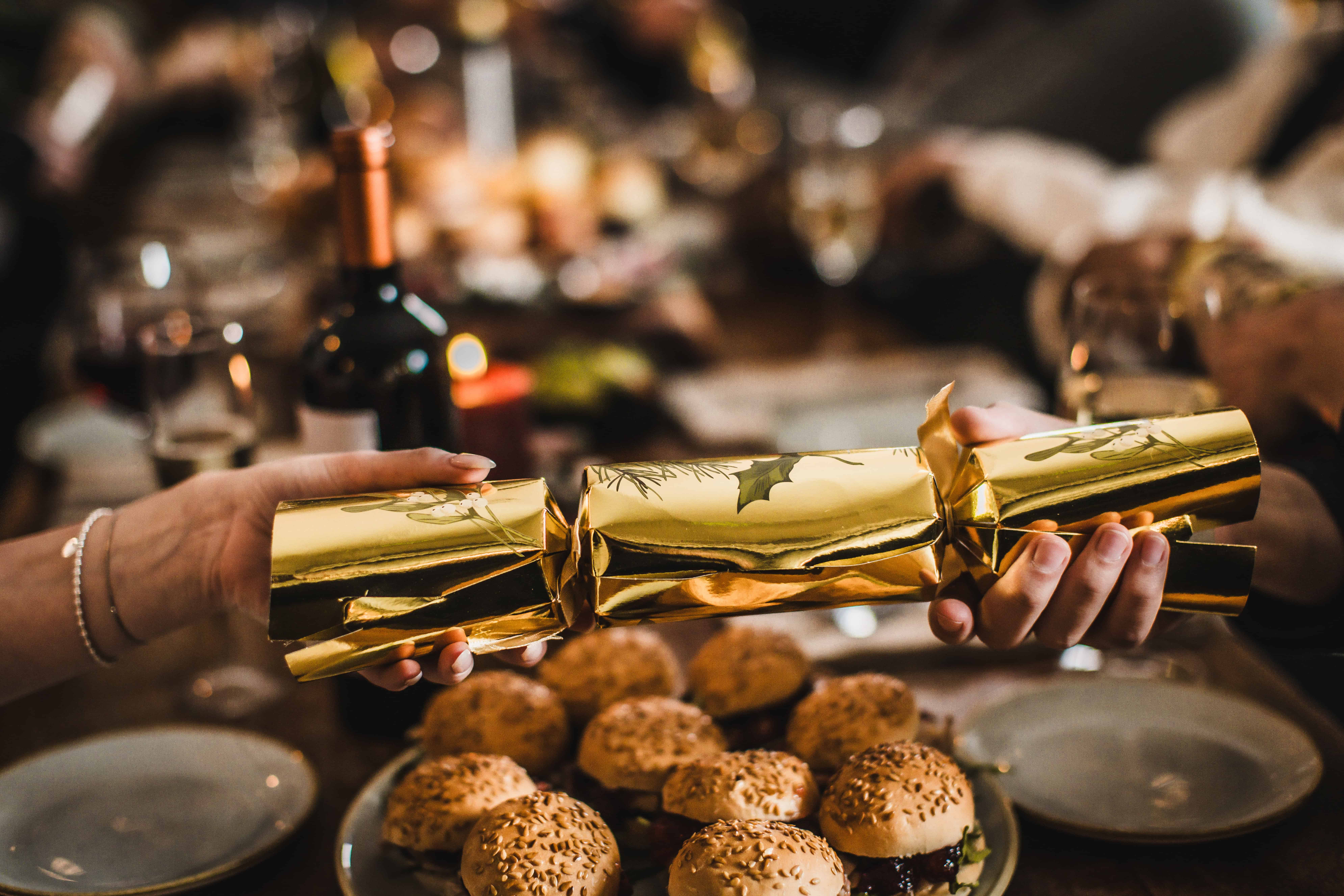 Hands holding a gold-wrapped Christmas cracker at a festive table.