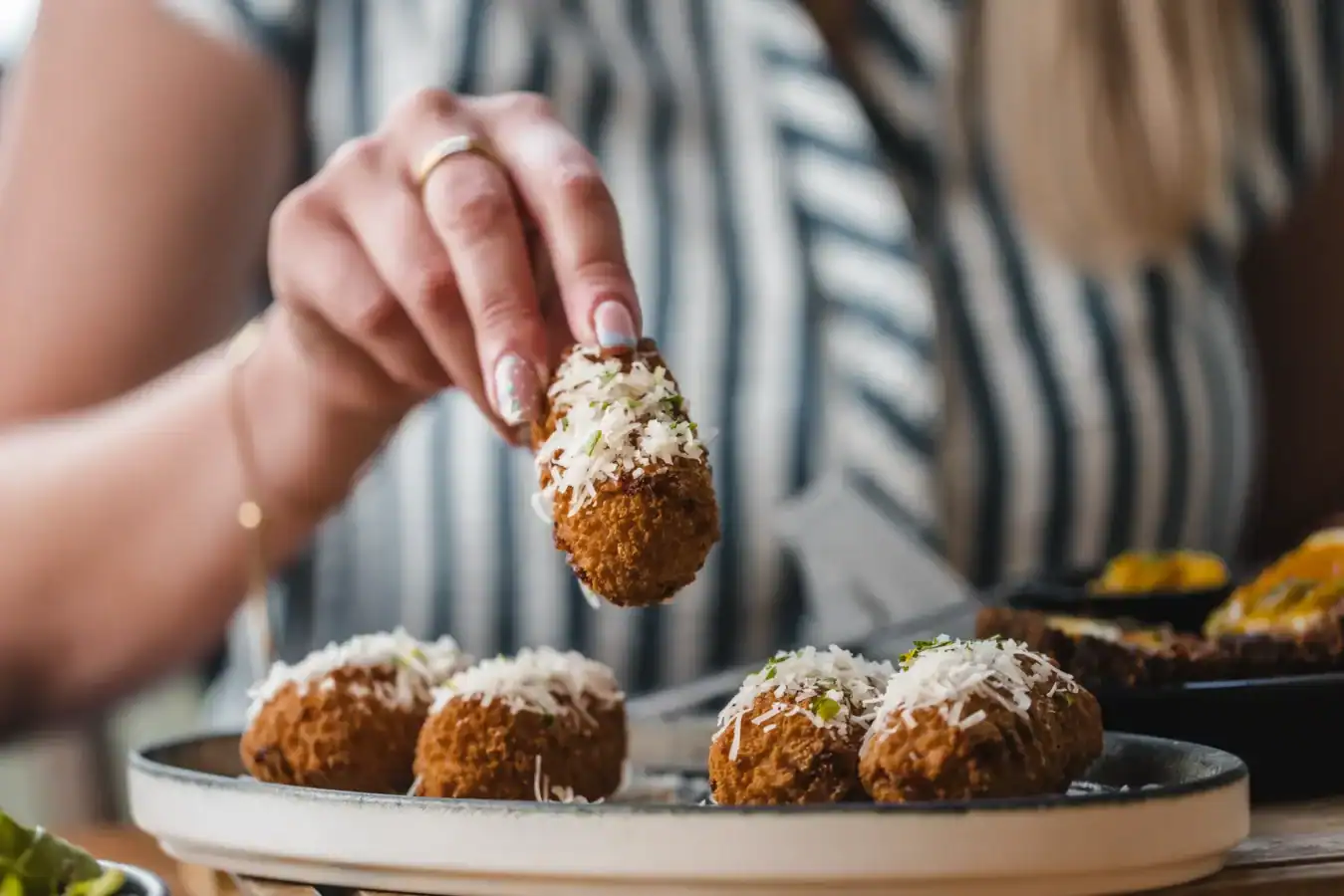 Close-up of hand holding a breaded appetizer with other snacks on a plate.