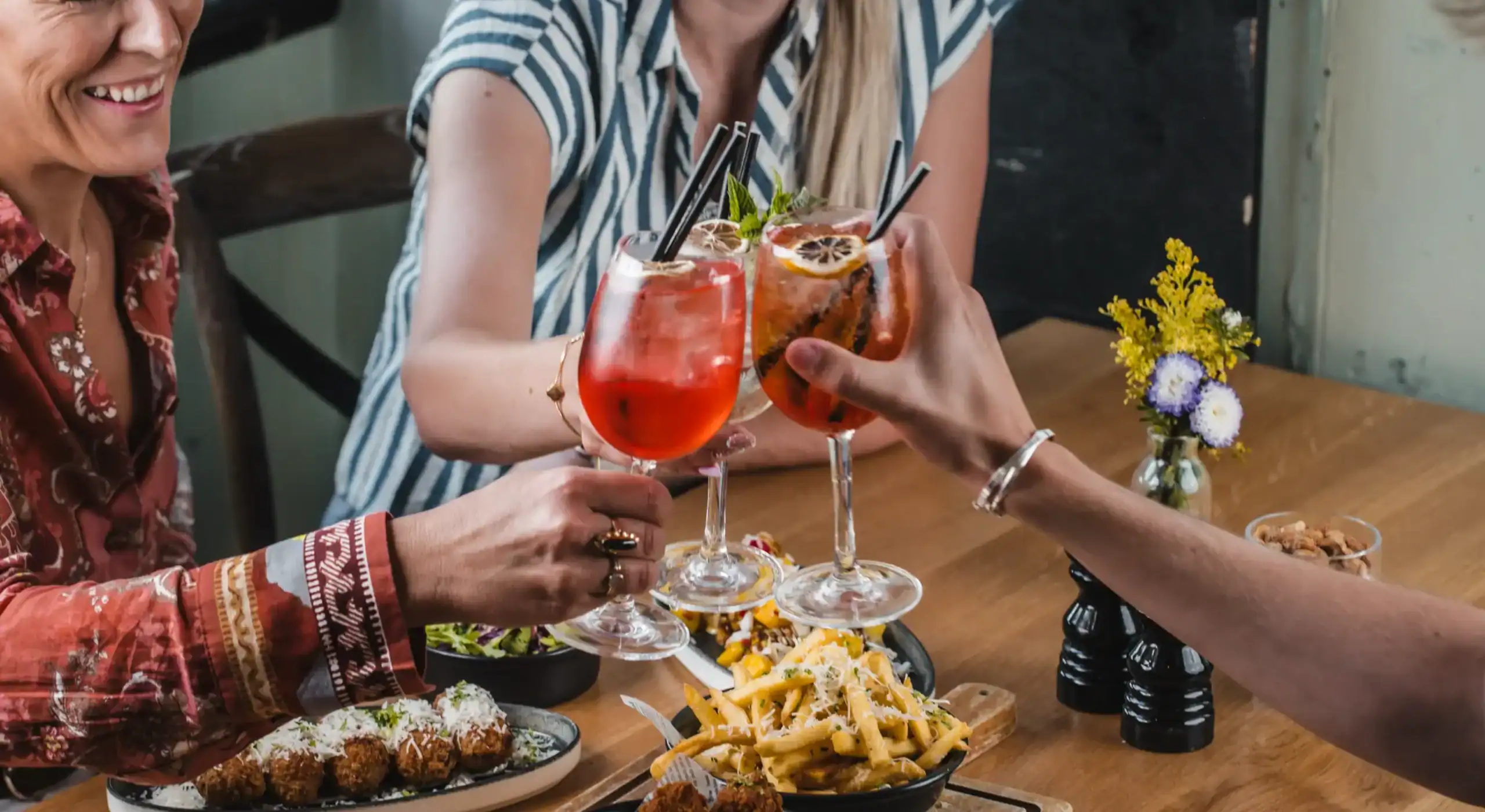 People clinking colourful cocktails at a restaurant table.